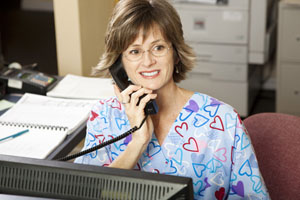 Medical administrative assistant wearing scrubs, talking on the phone behind a computer.