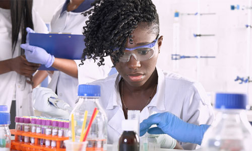 A young girl is sitting in a lab setting. She is wearing googles and a white lab coat while looking at a table full of equipment.