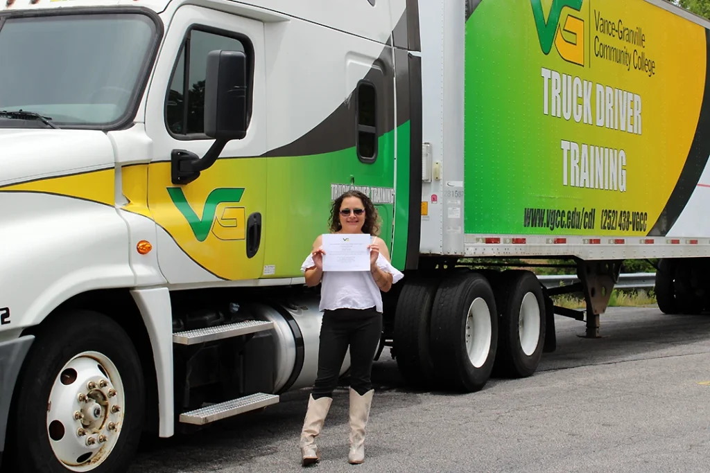 A female CDL Graduate holding a certificate in front of Vance-Granville's Truck Driver Training rig, which is painted green and yellow and displays the college's logo.