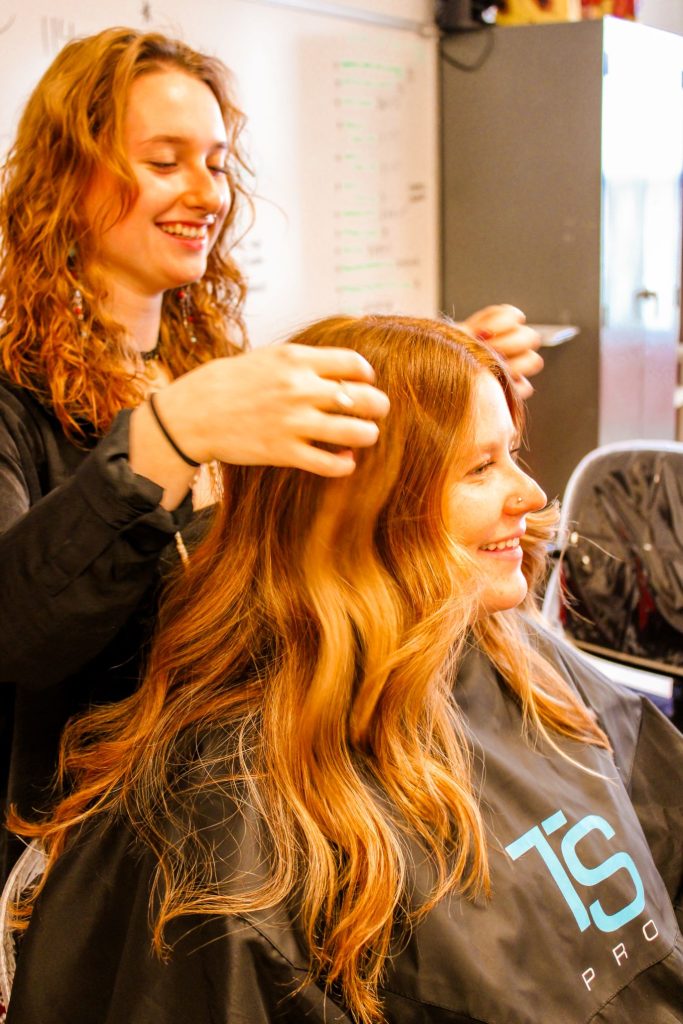 A young woman is smiling while working on another young woman's hair.