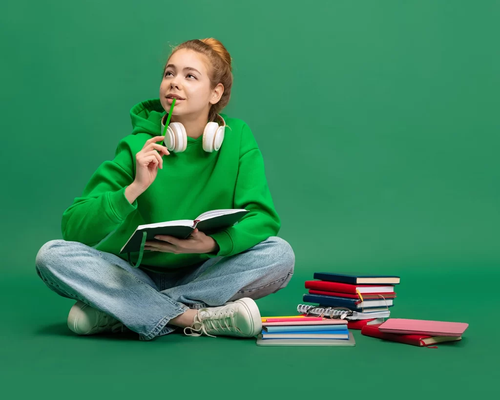Portrait of young girl, student in casual cloth sitting on floor with thoughtful expression, studying isolated over green studio background. [Shutterstock 2163185641]