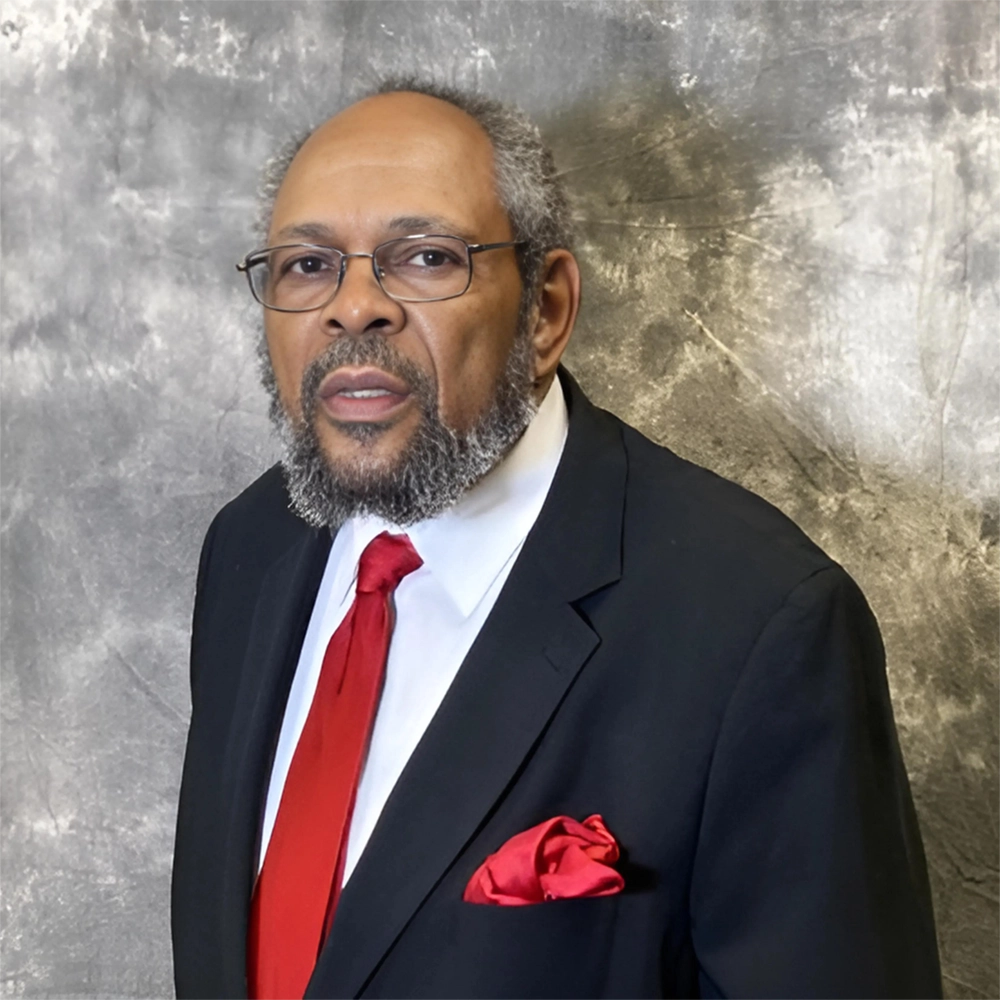 A formal portrait of a mature African-American man wearing a black suit, red tie, and red pocket square in front of a gray backdrop