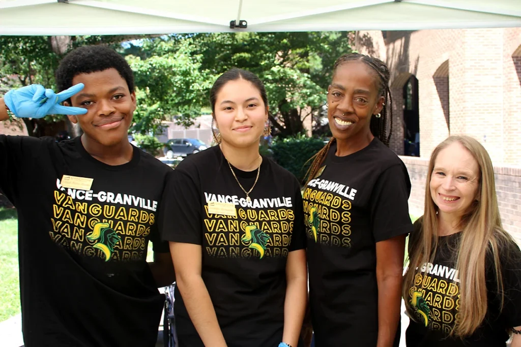 Four diverse college students wearing black T-shirts reading 'Vance-Granville Vanguards' while wearing blue rubber gloves to serve food outdoors.