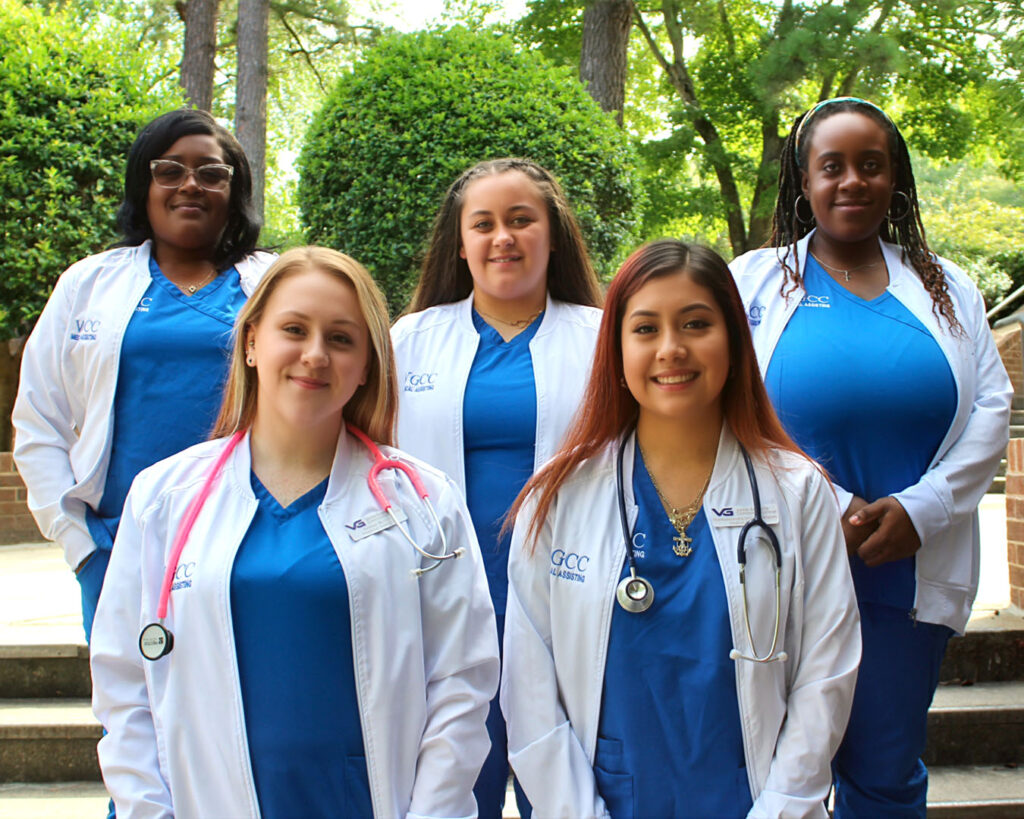 Five Medical Assisting graduates wearing blue scrubs and white medical coats