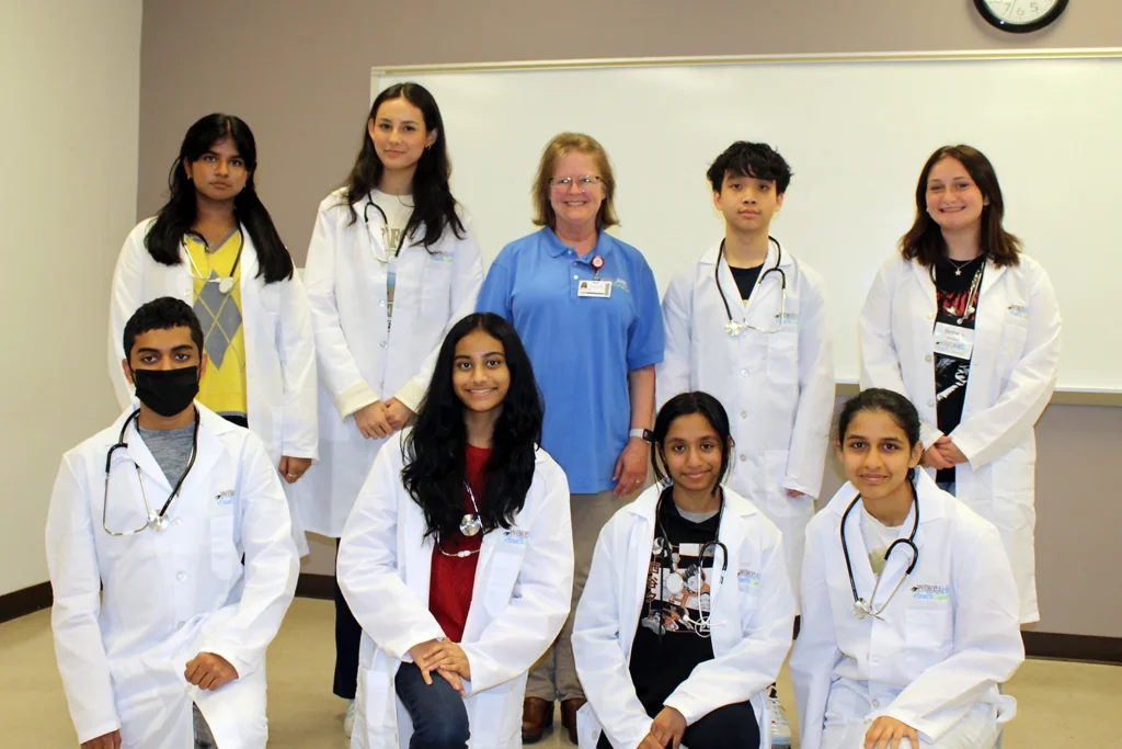 A uniformed woman stands in the middle of eight teenage students wearing lab coats and stethoscopes