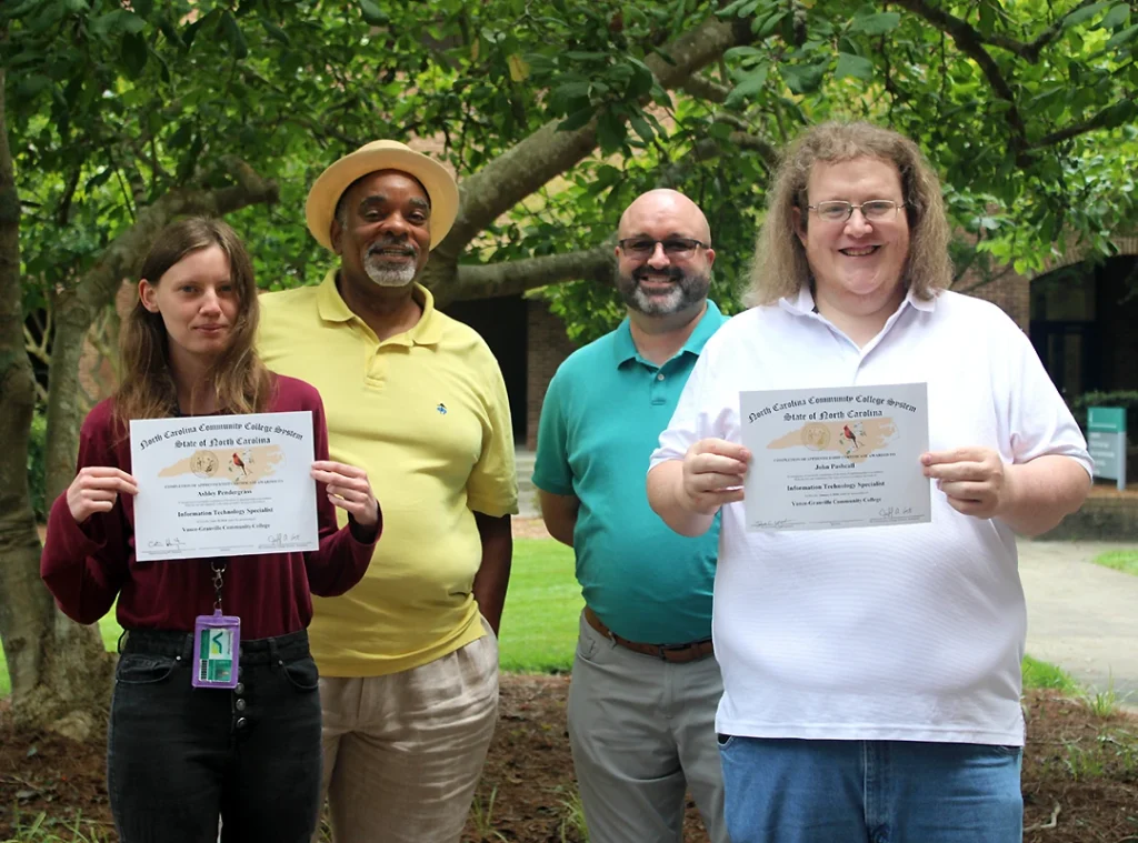 A young woman and young man hold apprenticeship completion certificates, while standing with two men who guided their apprenticeships.