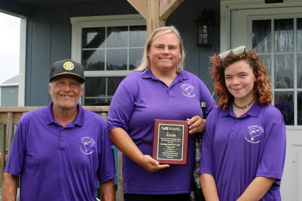 A family comprised of a man, woman, and teenage girl wear matching branded polo shirts. They stand on the exterior steps of a small building while the woman holds an award plaque.