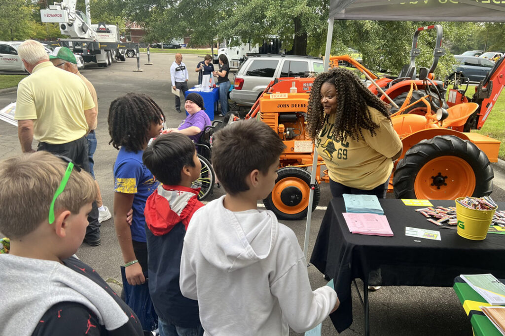 A woman in a yellow VGCC T-shirt, with an attentive expression on her face, leans toward a small group of middle school boys; other people and industrial vehicles are visible in the background.