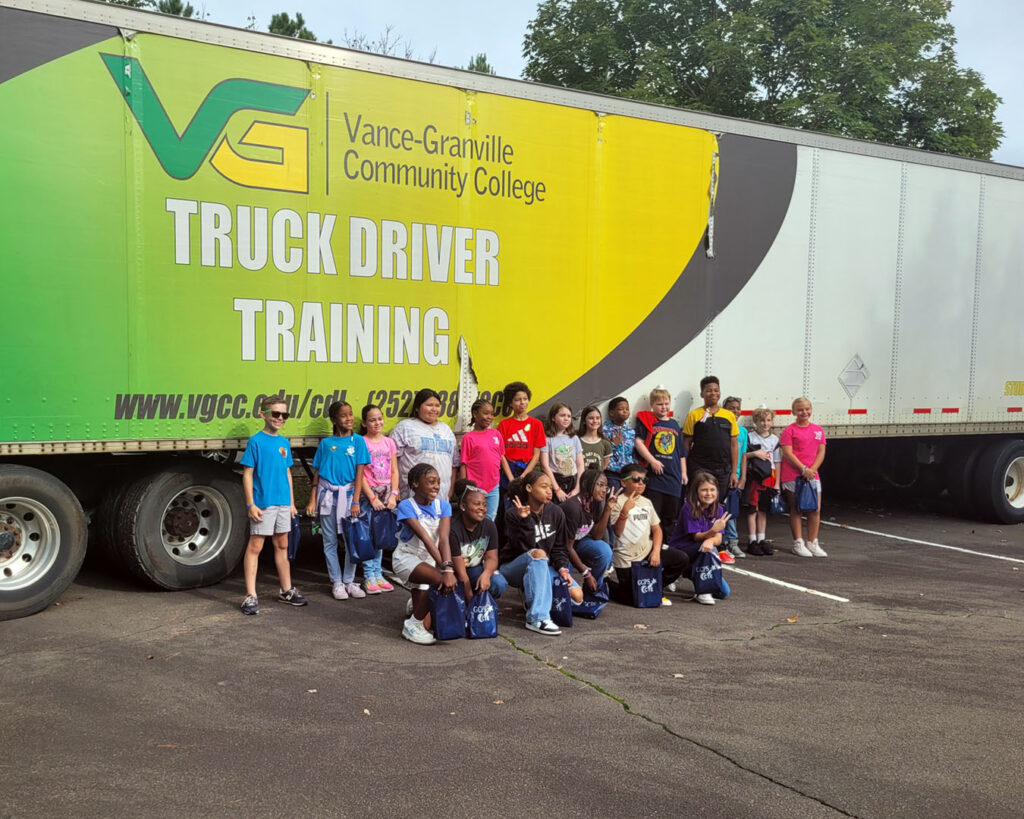 A group of about 20 middle school students poses for a group photo in front of a trailer from Vance-Granville Community College's Truck Driver Training program, which is painted green and yellow and displays the college's logo.