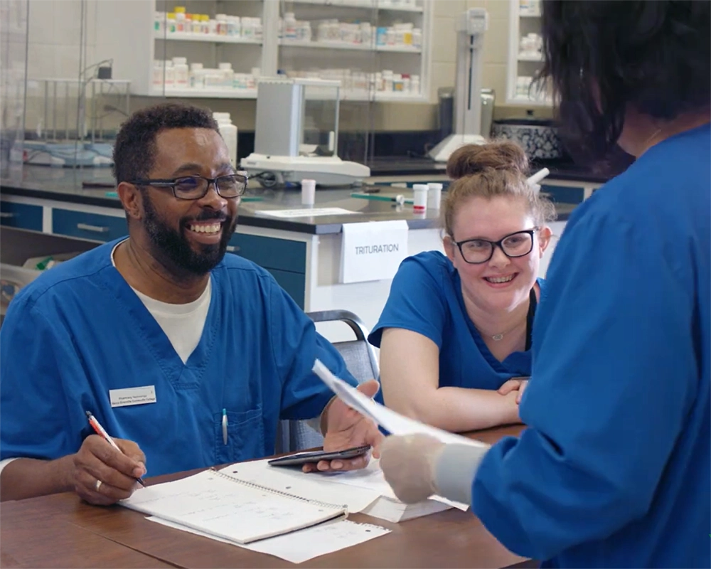 Two Pharmacy Technology students in blue scrubs, one male and one female, sit at a table in a lab and listen to an instructor who is not fully visible (video capture)