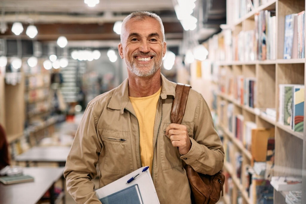An older man standing in the middle of a library with a bookbag and a notebook.