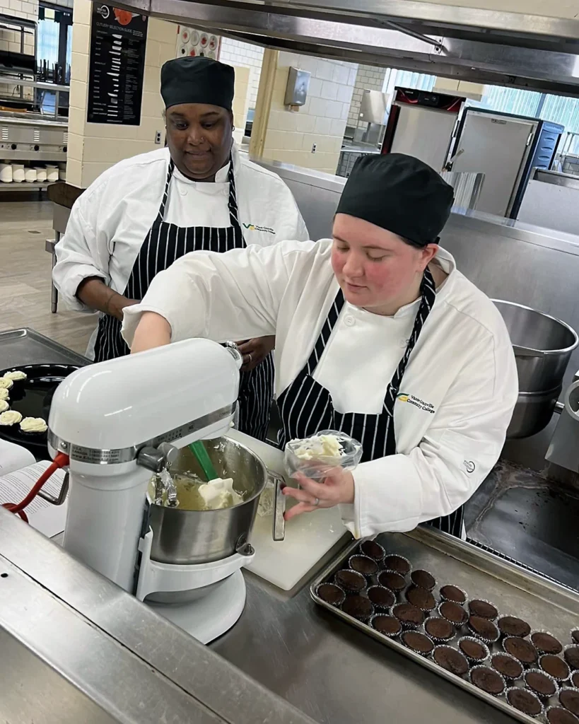 Two female culinary students prepare bite-sized desserts; one watches as the other scoops whipped topping from a stand mixer into a piping bag, preparing to pipe the topping onto a nearby tray of small chocolate desserts.