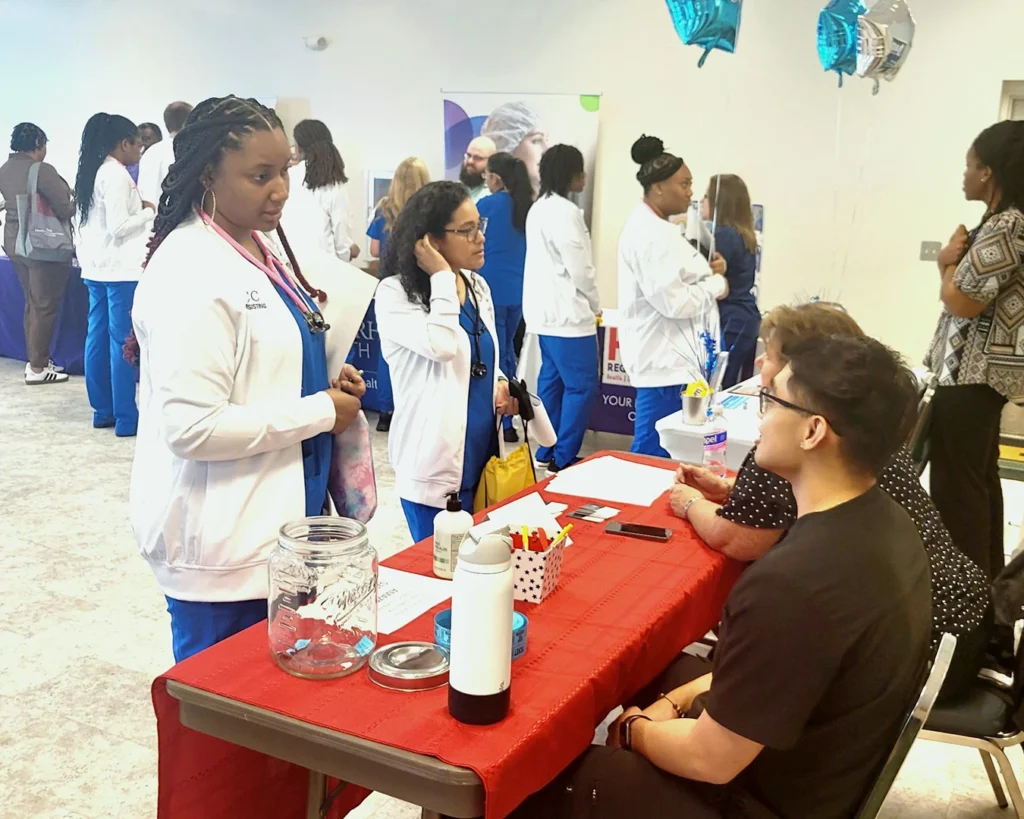 Students in blue scrubs and white medical coats stand at a vendor table and talk to seated job fair representatives; in the background, other students and members of the community visit other tables.