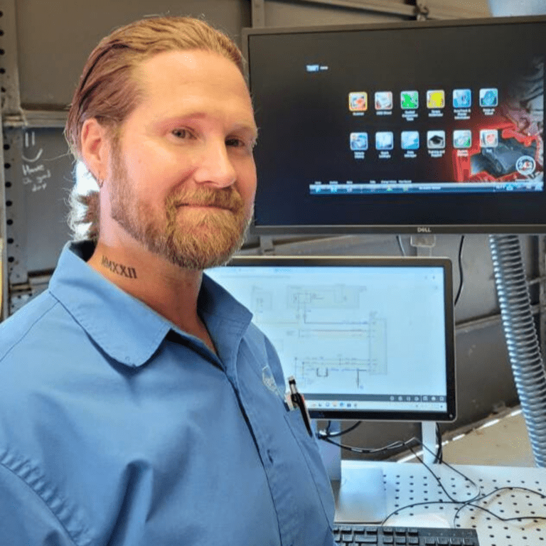 A man in a worker's jumpsuit stands in front of an electrical systems monitor [2025 Dallas Herring Award]