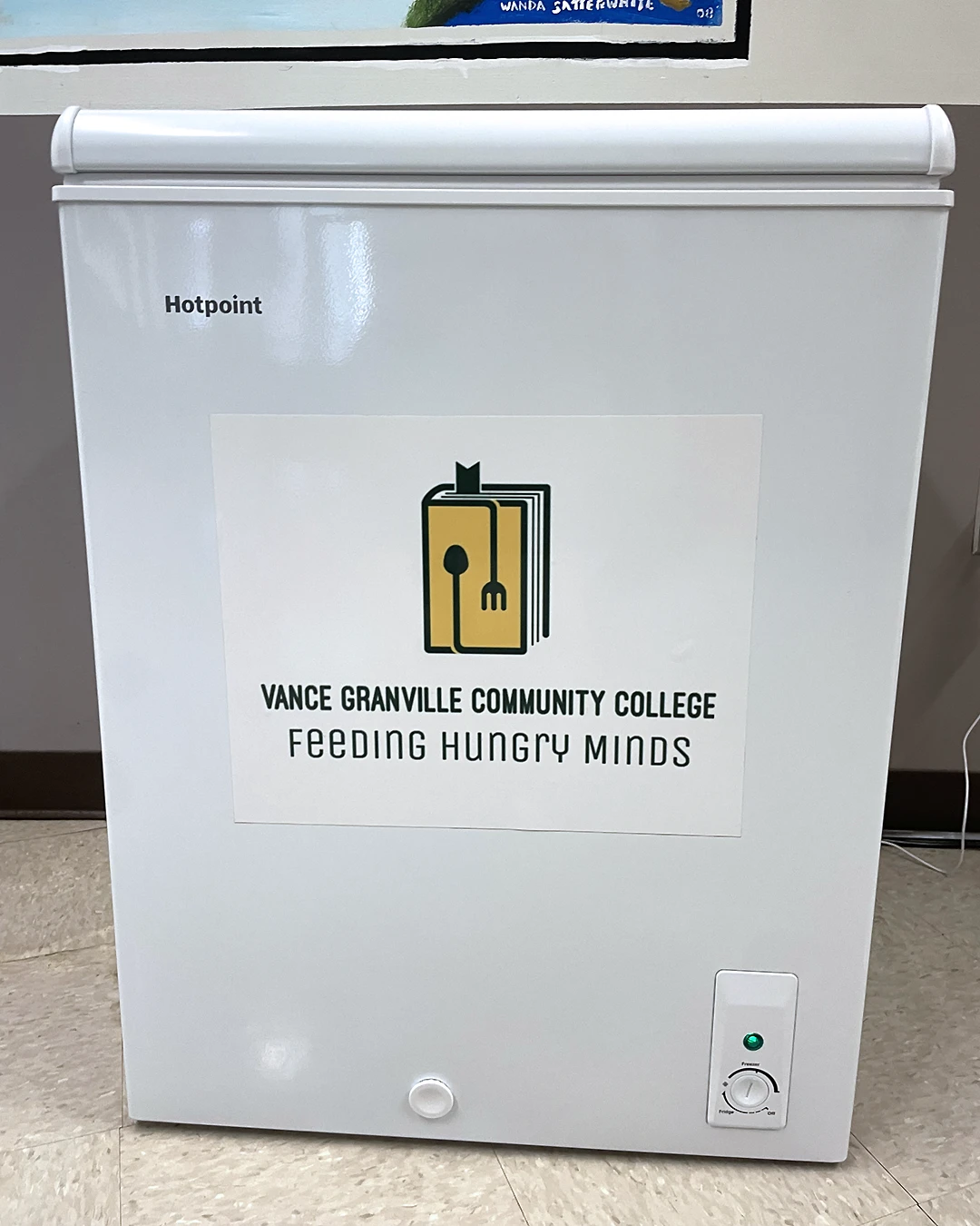 Closeup photo of a small freezer with a sign that reads 'Vance-Granville Community College Feeding Hungry Minds' beneath a graphic illustration that combines a fork and spoon with a book.
