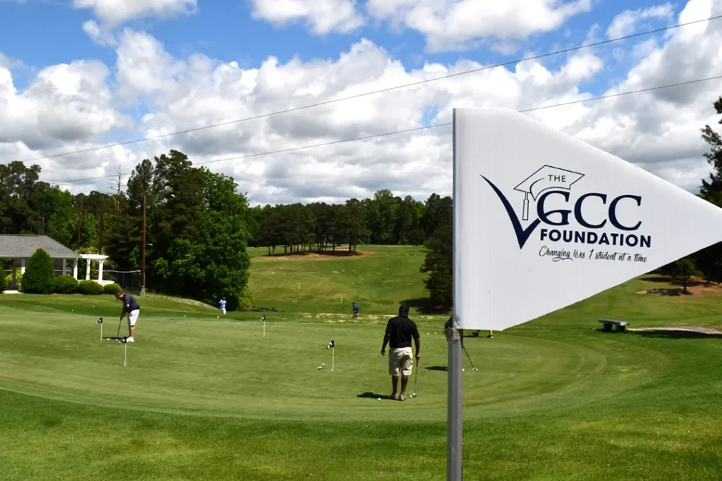 A golf flag branded with the VGCC Foundation logo is in the foreground; in the distance, a man putts during the 2024 VGCCF golf tournament.