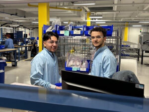 Two male-presenting apprentices wear safety glasses and smocks while working at an industrial facility