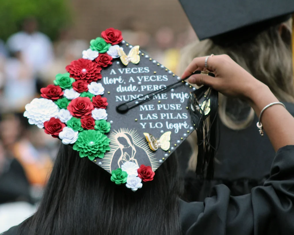 Closeup photo of a graduate's embellished cap. It's decorated with green, white, and red flowers; gold accents; and Spanish text that's translated 'Sometimes I cried, sometimes I doubted, but I never gave up. I got my act together and I made it.' [2024 commencement]
