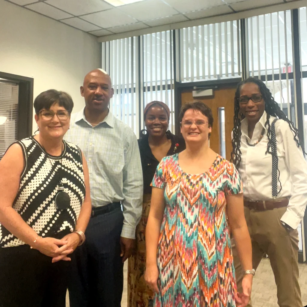 Four diverse VGCC employees pose with recent graduate Nikita Rodebaugh, in a college office with large windows and vertical blinds behind them.