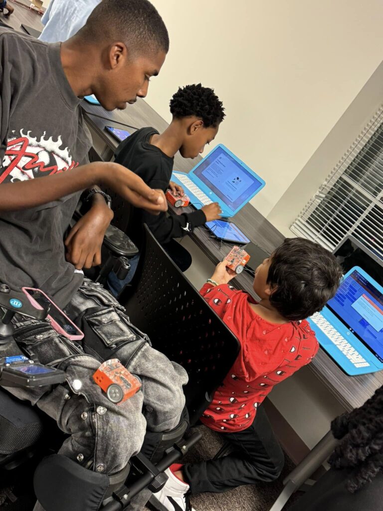 A man in a motorized wheelchair engages with two children, who are using laptops to program small wheeled robots.
