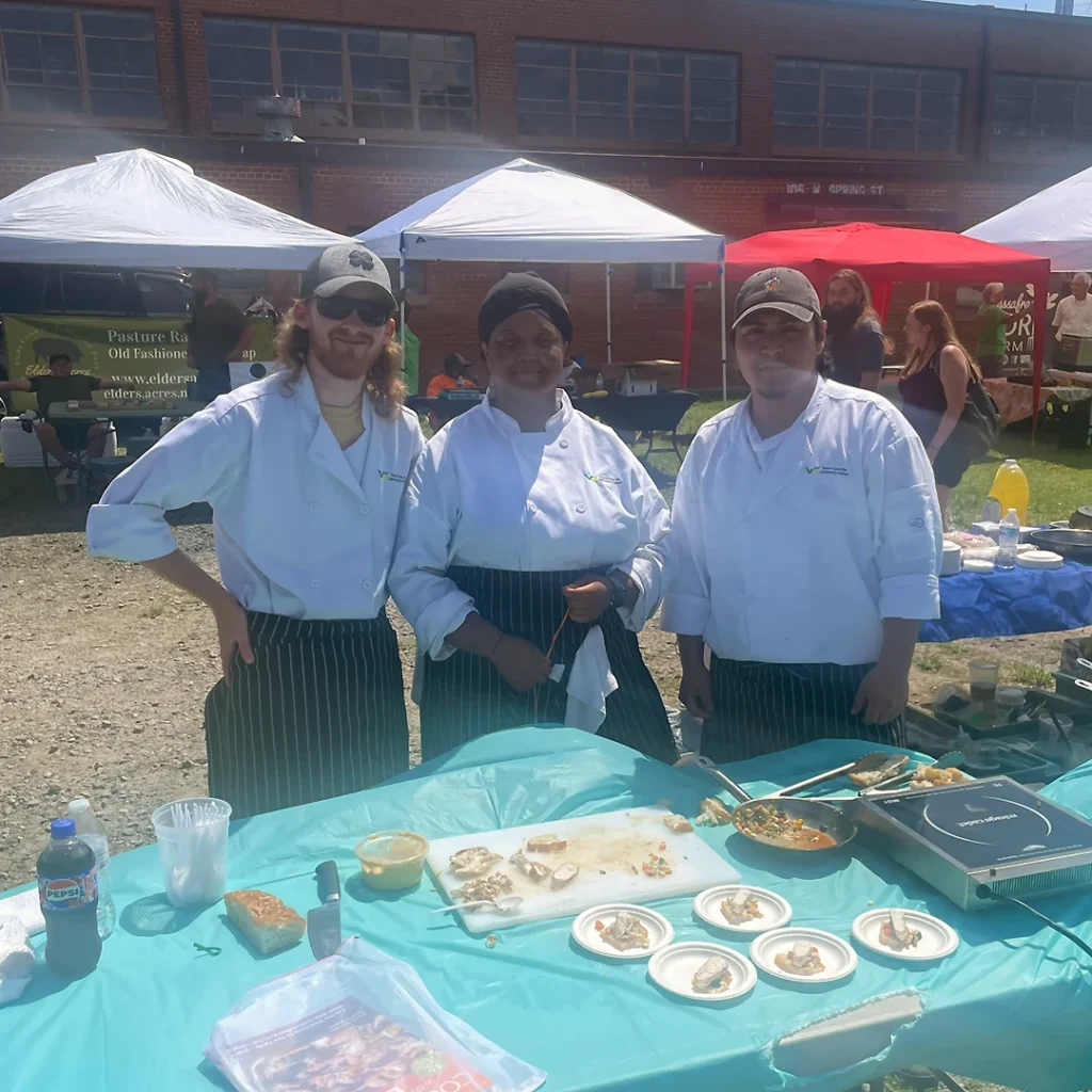 Three diverse culinary students in chef's coats and aprons pose together during a cooking competition at a bustling, sunny, outdoor farmers market.