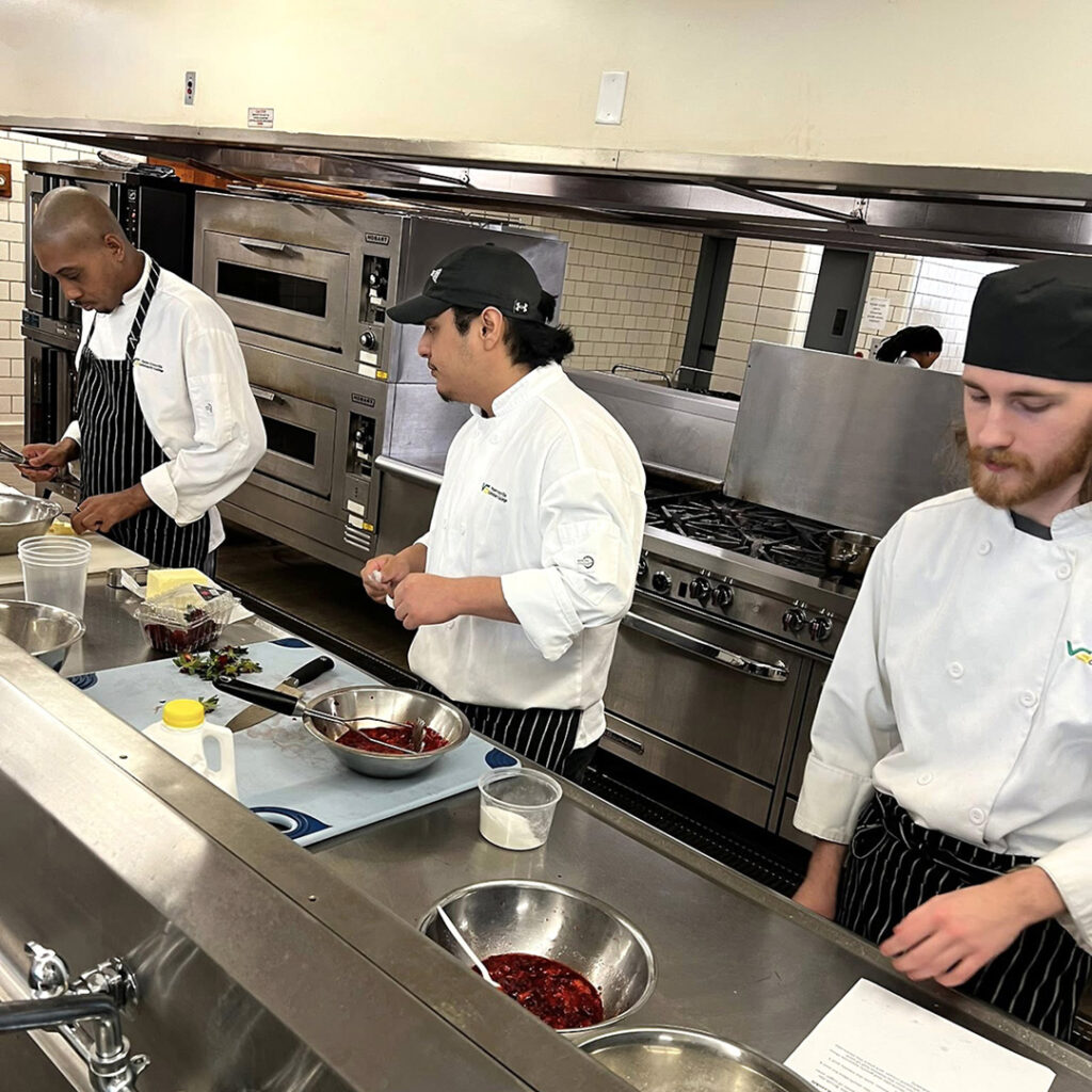 Three male Culinary Arts students use knives, cutting boards, and bowls to prepare a dish with strawberries in an industrial kitchen.