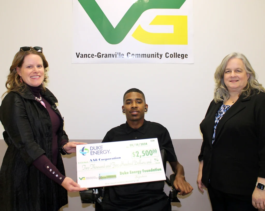 A smiling man in a motorized wheelchair sits between two women, who are presenting him with grant check for AA8 Corporation