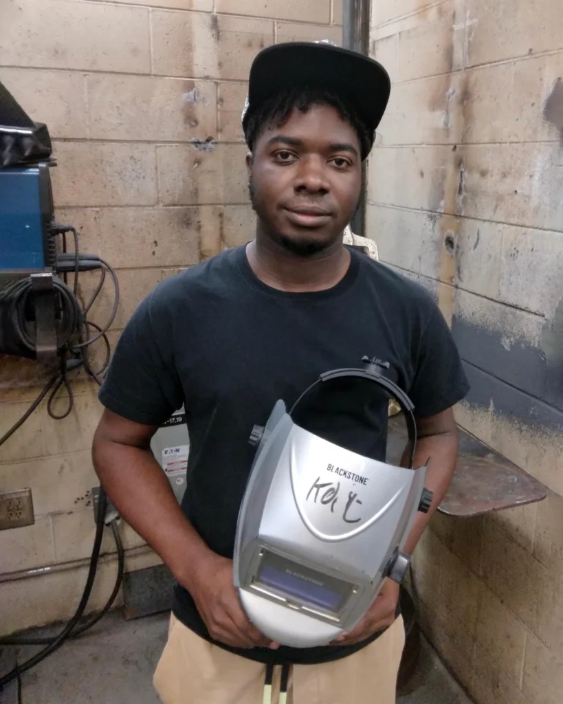 Kobe Bullock, a young Black man, stands in a welding workshop and holds a safety helmet bearing his signature.