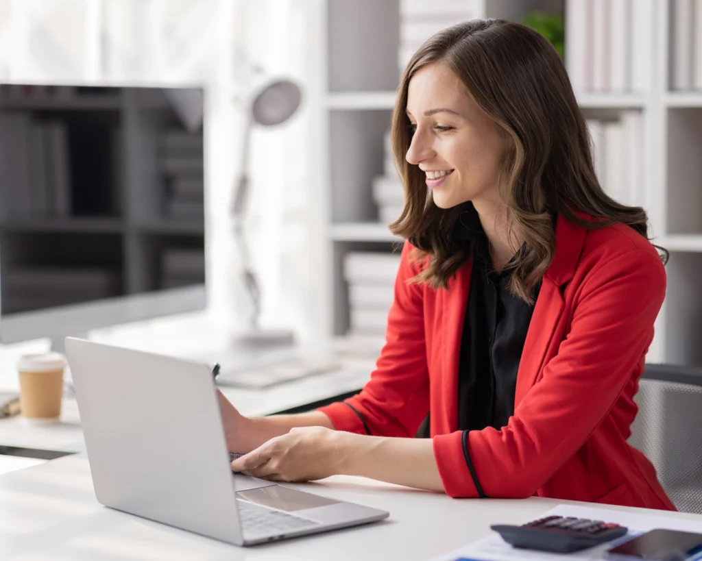 Professional young woman in a red jacket sitting at desk working on laptop [administrative careers]