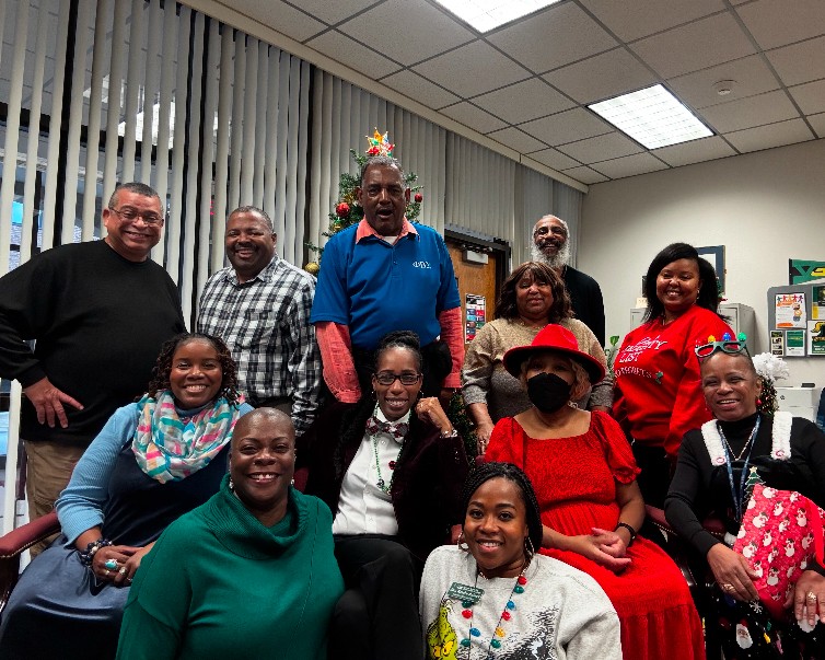 Group of twelve people, dressed in casual and festive attire, gathered in an office with a decorated Christmas tree in the background.
