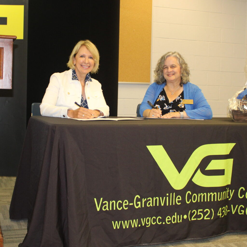 Two women in professional attire sit at a table covered in a Vance-Granville Community College tablecloth and sign papers formalizing VGCC's transfer agreement with Northwood University