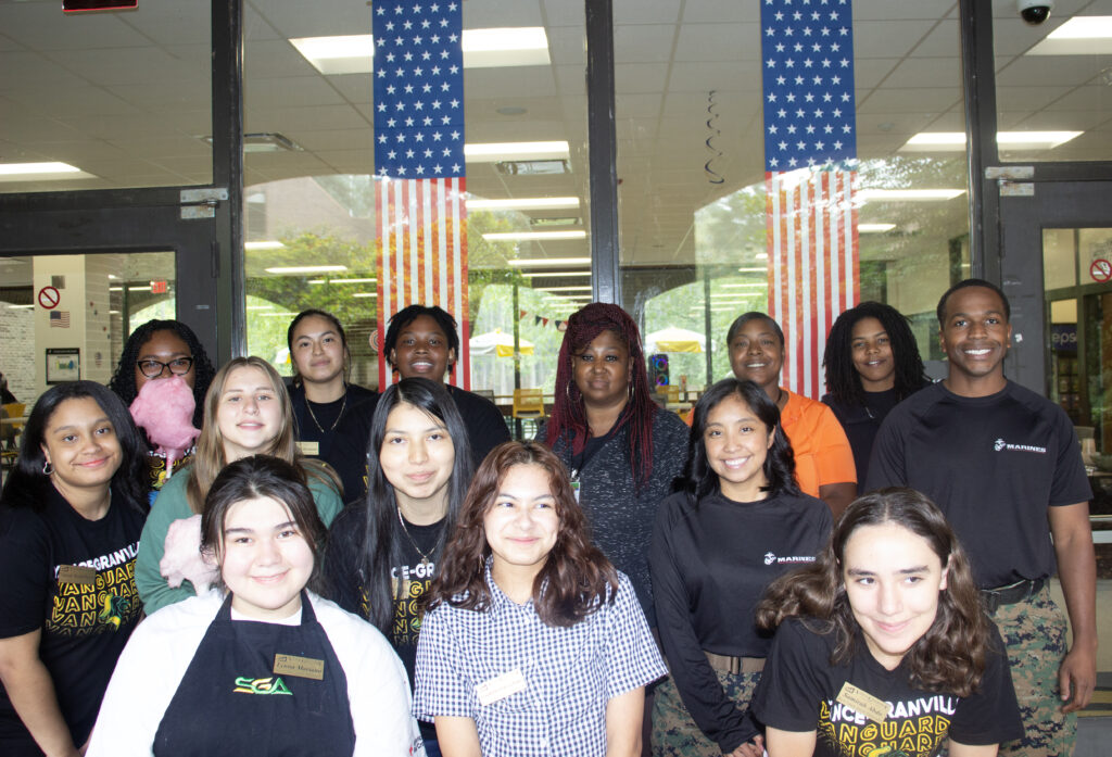Approximately fourteen VGCC students stand smiling in front of the student lounge, one of whom is holding Cotton Candy. Flag decals hang in the windows behind them.