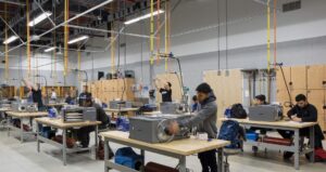Students work on individual HVAC units that sit upon industrial style work benches. Lockers line the back walls while orange and yellow pipes extend from the ceiling over ever work bench. This demonstrates how the future HVAC lab in the new Center for Advanced Manufacturing and Technology may look.