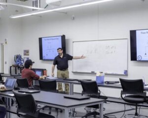 A teacher stands in front of long tables that have laptops in front of each chair. Two TVs hang on either side of a white board. This is how a classroom in the new Center for Advanced Manufacturing and Technology may look.