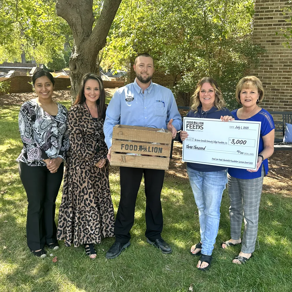 Four female employees of Vance-Granville Community College and a male Food Lion representative stand on a grassy lawn, holding a large presentation check from the Food Lion Feeds Foundation