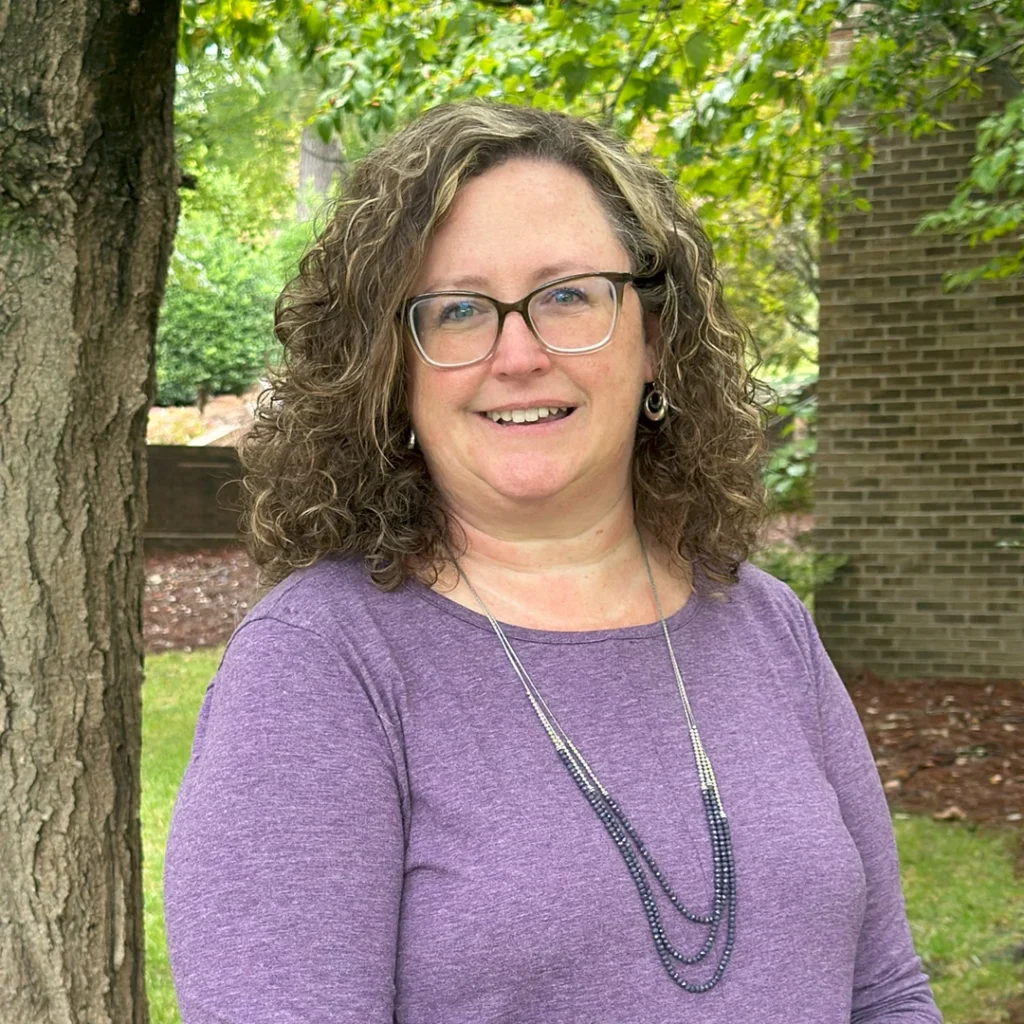 Headshot of Marsha Overby, a smiling middle-aged woman wearing a purple top and necklace while standing under a leafy green tree.