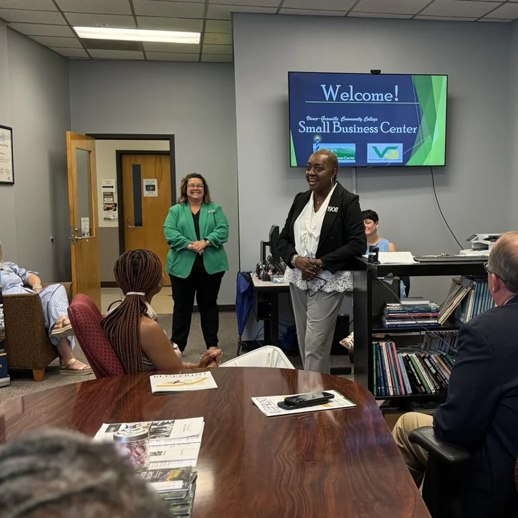 A professionally dressed woman speaks to guests seated around a shiny wooden table; a digital sign behind her reads 'Welcome! Vance-Granville Community College Small Business Center.'