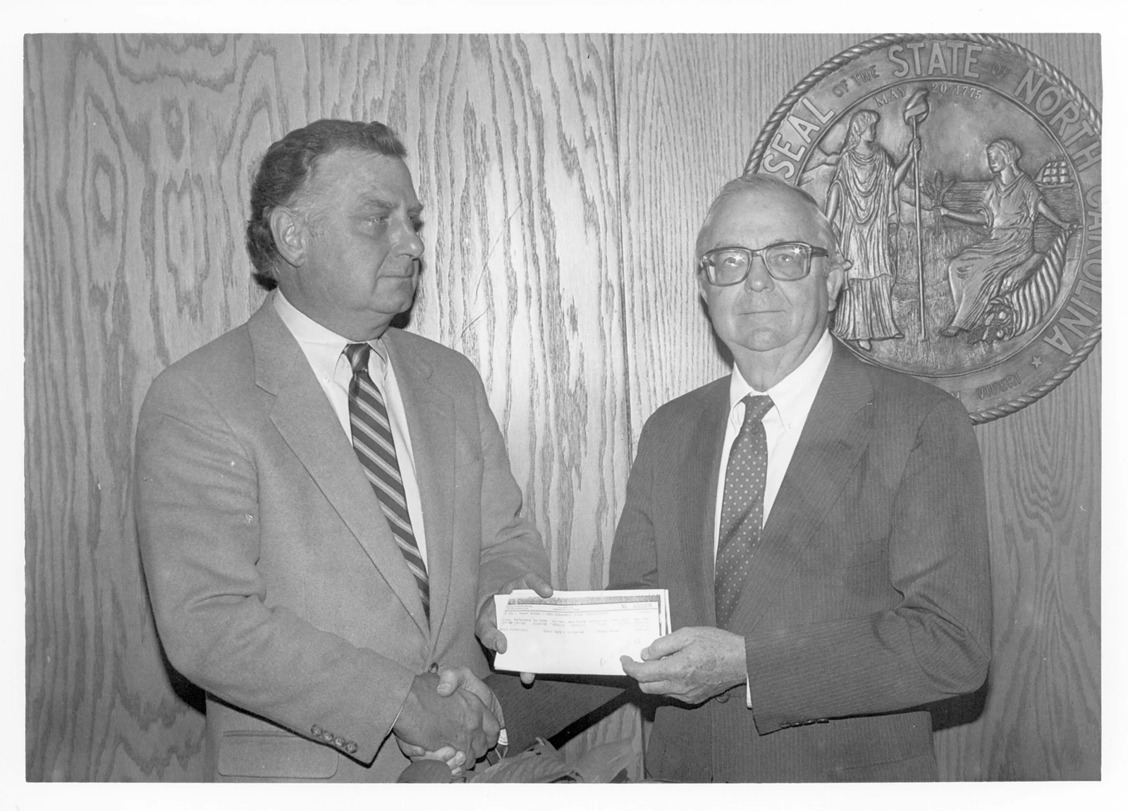 A black and white photo of two men in business suits, holding a donation check while standing in front of a large North Carolina state seal.