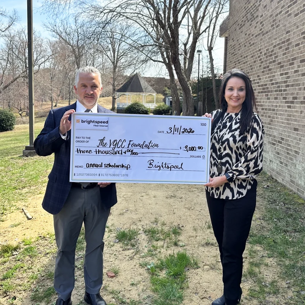Two individuals in business attire stand outdoors on the VGCC Henderson campus, holding a large presentation check representing a $3000 donation.