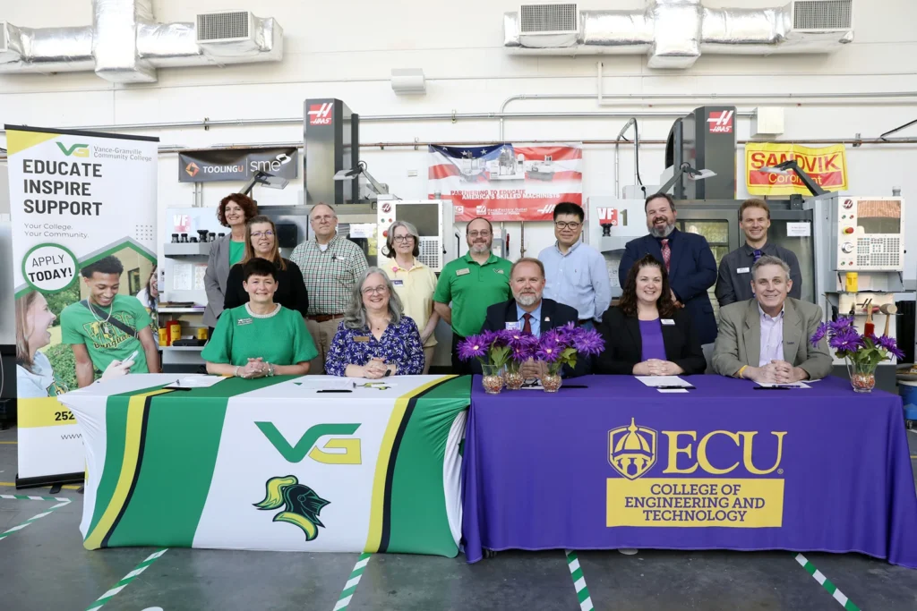 A group of 13 people representing the administration and faculty of Vance-Granville Community College and East Carolina University gather behind tables bearing their respective institutions' branding, inside a large ECU engineering lab.