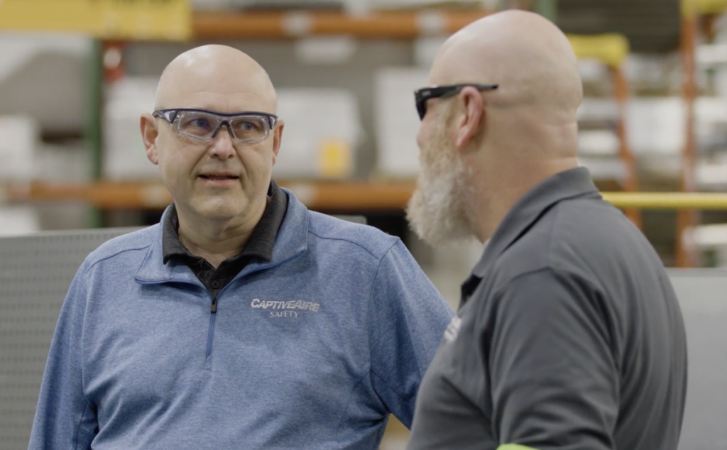 Chris Clough from Captive Aire stands with another person inside an industrial looking room. Both are wearing safety glasses.