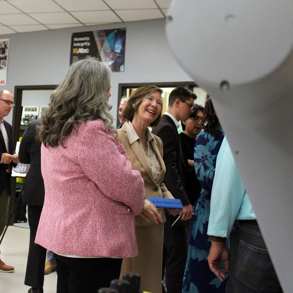 Two professionally dressed women engage in conversation in a college mechatronics lab while surrounded by robotics equipment and a diverse crowd of people.