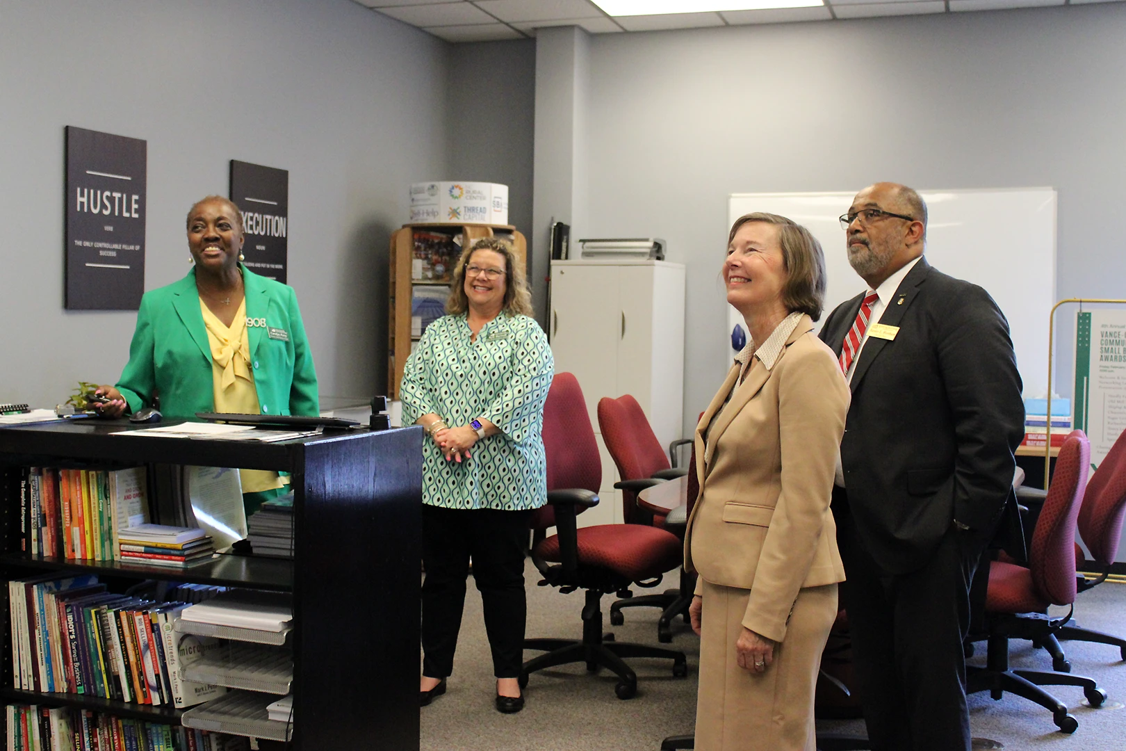 Four adults in professional clothing stand in a conference-style room surrounded by bookshelves and inspirational posters; they all look left to watch a digital screen just outside of the photo's frame.
