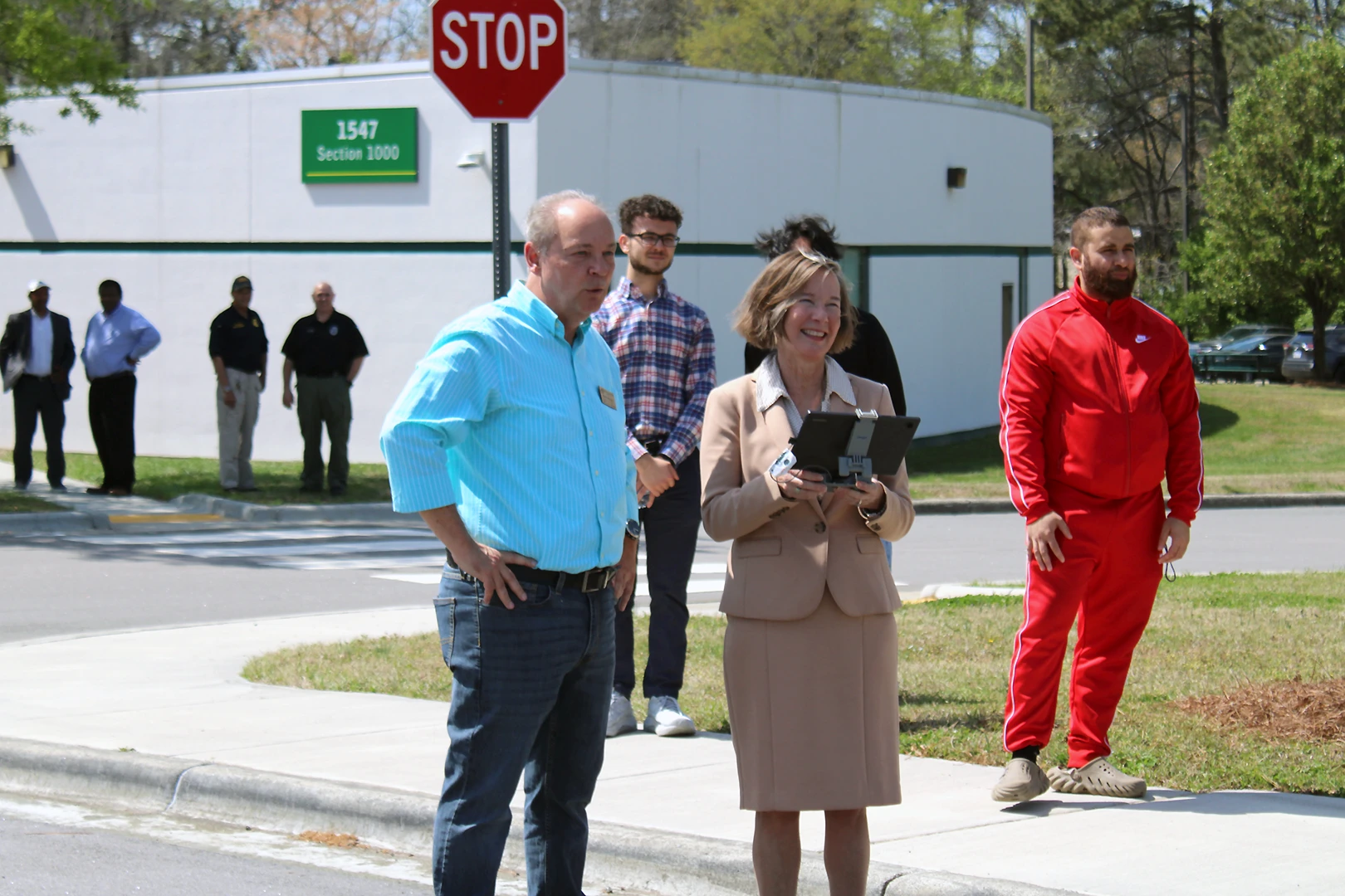 A man in business casual attire instructs a woman in a skirt suit how to operate a drone using a touchscreen tablet; they stand in the parking lot of VGCC's South Campus as students and college representatives watch from the background.