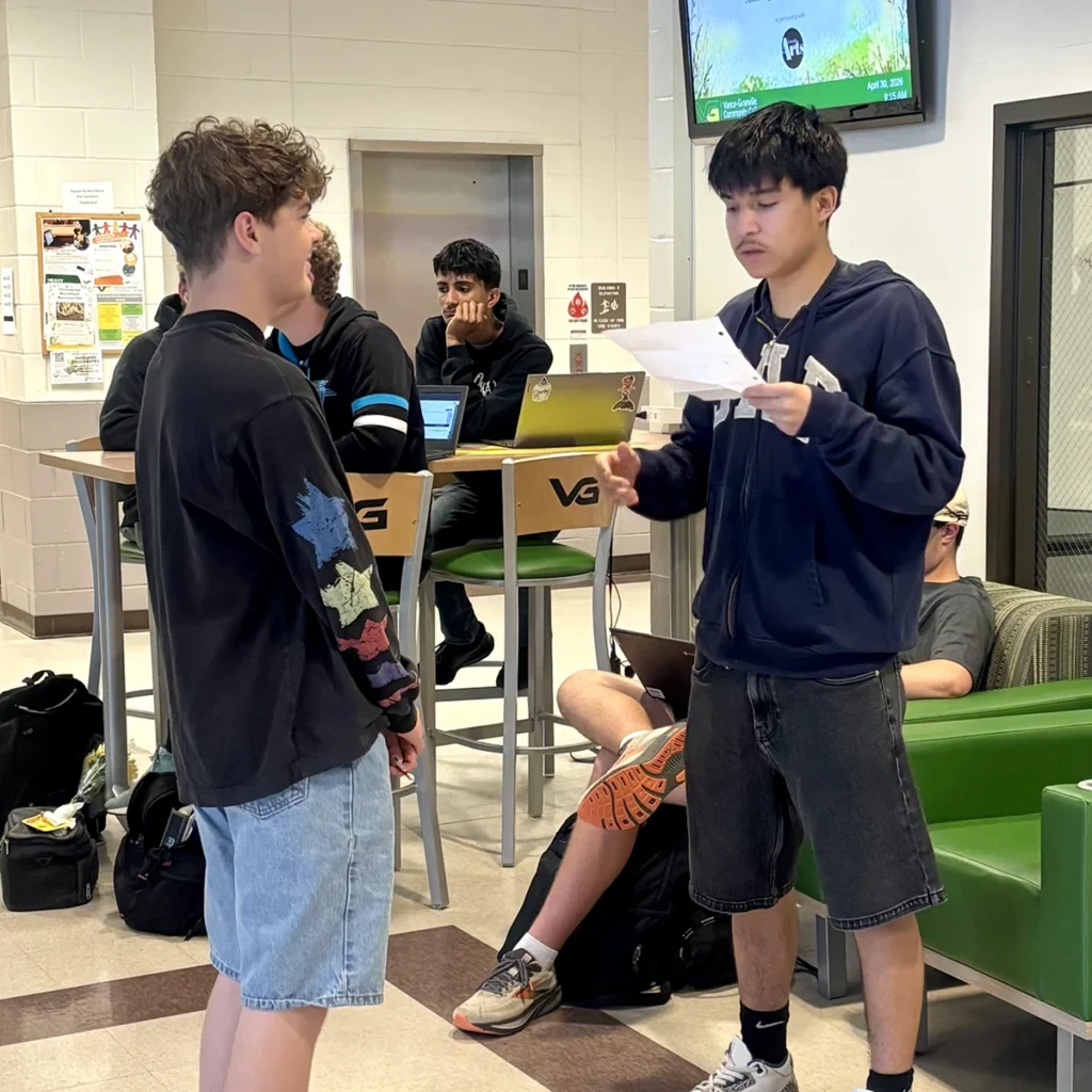 A college student stands in a bustling campus lounge and reads from a sheet of paper as his peers listen.
