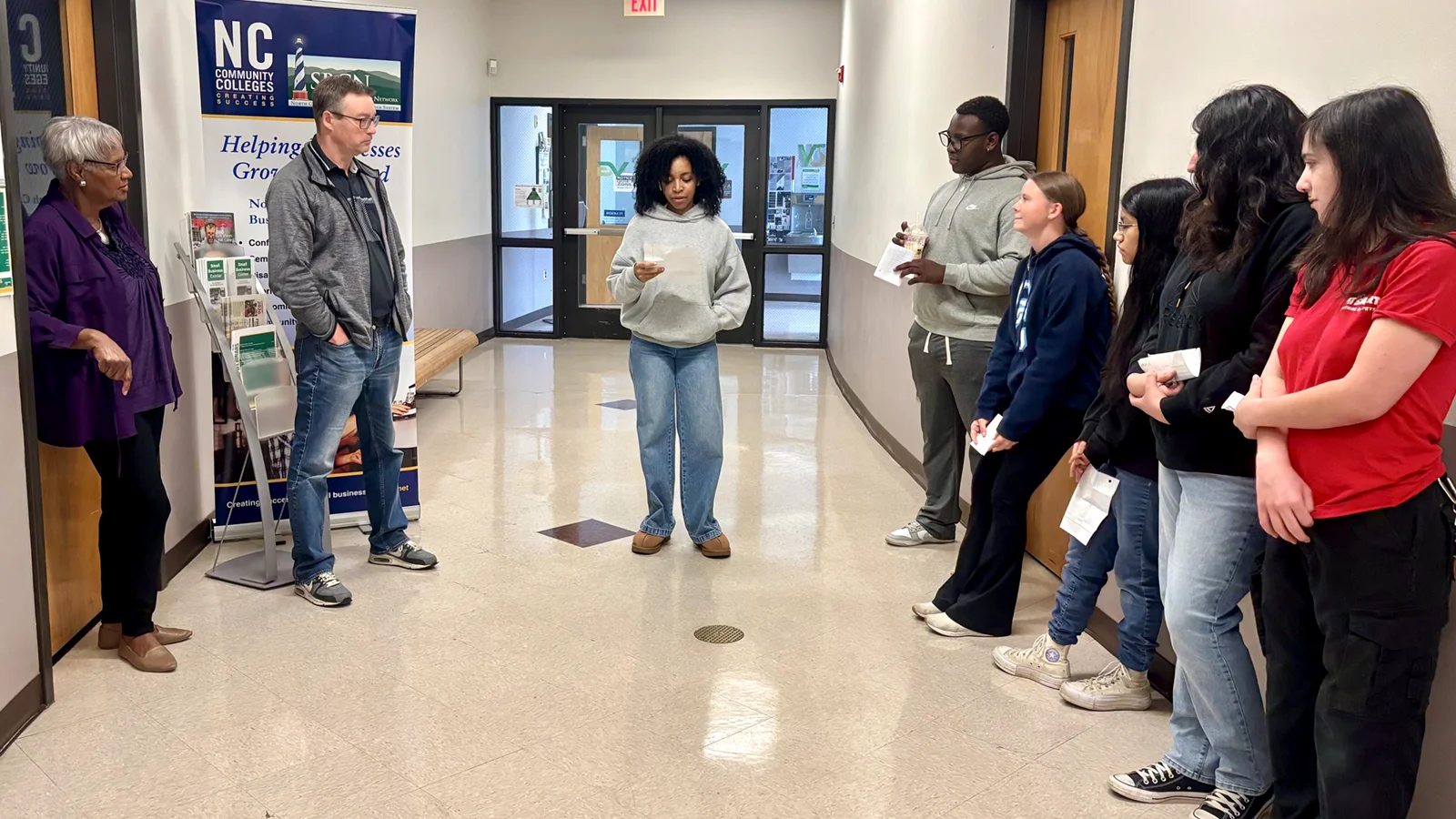 Five young adult students and two college employees stand listening in a hallway as another student reads from a sheet of paper.