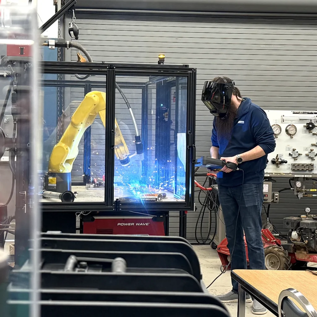 An individual wearing a black welding hood uses a handheld device to operate a yellow machine, which is giving off sparks behind protective glass.