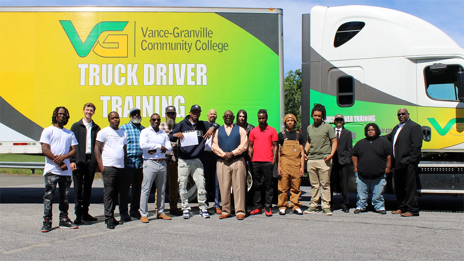 16 diverse adults pose in front of a large tractor trailer, which is covered in vibrant green and yellow graphics, the Vance-Granville Community College logo, and text reading 'Truck Driver Training.'