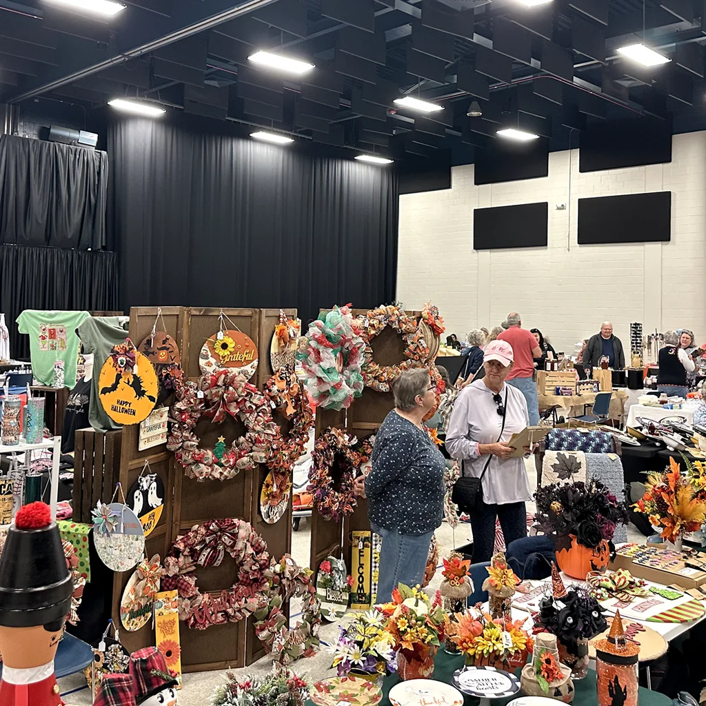Rows of tables and standing displays covered with handmade goods inside a large multipurpose room, with people talking and browsing.