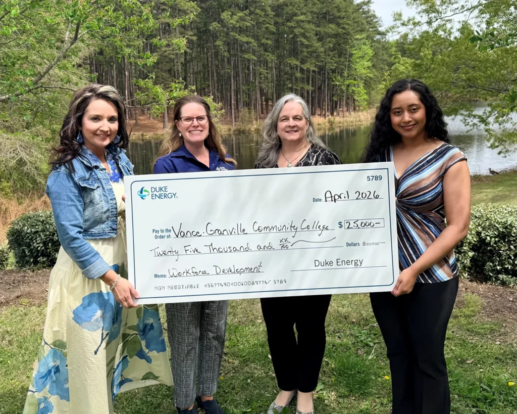 Four women hold a large presentation check in front of a pond surrounded by green leafy trees.